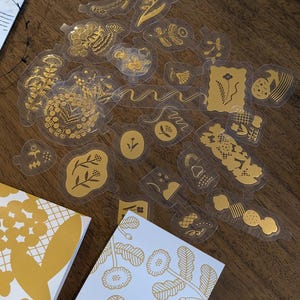 May include: A collection of gold-colored stickers with various floral and abstract designs, scattered on a wooden surface. Two notebooks with gold floral patterns are also visible. The stickers appear to be transparent with gold foil details.
