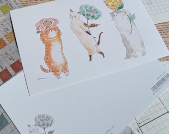 May include: Two postcards featuring watercolor illustrations of cats holding flowers. One card shows three cats with different colored fur, each balancing a flower. The second card displays a single cat with a flower. The cards are on a patterned background.