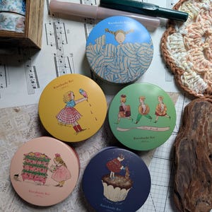 May include: A collection of round tins with illustrations, featuring a girl with an ice cream cone, a girl with a cupcake, and other whimsical designs. The tins are in various colors, including blue, yellow, and pink. Also visible are pens, washi tape, and a wooden box.
