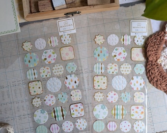May include: Two sheets of decorative stickers with various shapes, including circles, squares, and hexagons. The stickers feature pastel colors and patterns like polka dots and floral designs. The brand name "WANOWA" is visible on the packaging.