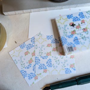 May include: Stationery set featuring floral patterned memo pads and cards. The design includes blue and pink flowers with green stems on a white background. A green fountain pen is also visible.
