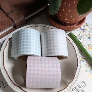 May include: Three rolls of washi tape with gingham patterns in light blue, beige, and pink. The rolls are on a white plate with a brown rim.