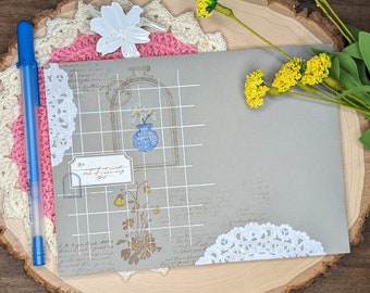 May include: A gray envelope with a white grid pattern and floral illustrations. The envelope has a white lace doily adhered to the bottom right corner. The envelope is on a wooden surface with a pink and white crocheted doily and a blue pen.