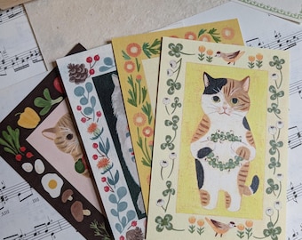 May include: A collection of illustrated cards featuring cats, flowers, and other nature-themed designs. The cards have decorative borders and are arranged on a background of sheet music. A wooden box of stamps and a small bowl of paper clips are also visible.