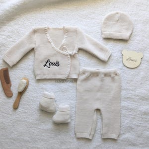 Peut inclure: Ensemble bébé en tricot blanc composé d'un cardigan, d'un pantalon, d'un bonnet et de chaussons. Le cardigan a une fermeture enroulée et est brodé du nom "Louis".