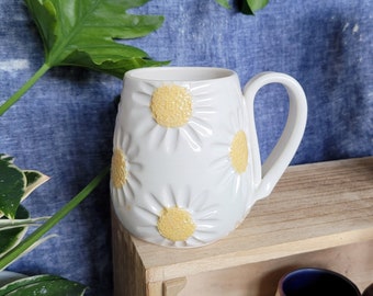 Puede incluir: Taza de cerámica blanca con un diseño de margarita en relieve. Las margaritas tienen centros amarillos y pétalos blancos. La taza tiene un asa y está sentada en un estante de madera.