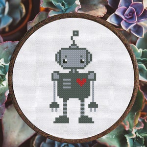 May include: A gray and red pixelated robot with a heart on its chest, embroidered on a white canvas in a round wooden frame.