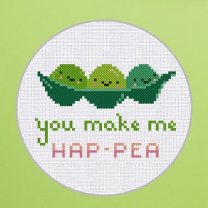 Può includere: Un ricamo a punto croce bianco con tre piselli verdi in un baccello. I piselli hanno facce felici e il testo "You make me hap-pea" è scritto sotto.