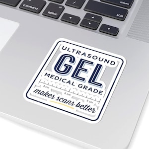 Op de afbeelding: Een witte sticker met een blauwe rand en de tekst "ULTRASOUND GEL MEDICAL GRADE" in een vet, marineblauwe lettertype. De sticker bevat ook de tekst "makes scans better" en een liniaal met metingen in centimeters.