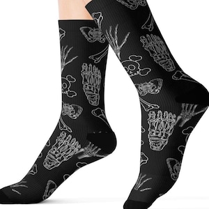 May include: Black socks with a white skeletal pattern. The design includes illustrations of bones, skulls, and hand and foot bones. The socks are worn on the feet.