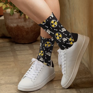 Puede incluir: Un par de calcetines negros con un patrón amarillo y negro de símbolos de radiación y huesos. Los calcetines se llevan con zapatillas blancas.
