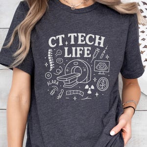 Könnte beinhalten: Dunkelgraues T-Shirt mit dem weißen Text "CT TECH LIFE" und medizinischen Grafiken. Das Design umfasst einen CT-Scanner, Wirbelsäule, Gehirn und andere medizinische Symbole. Der Name "ALARA" ist ebenfalls auf dem Shirt aufgedruckt.