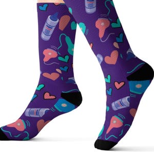 Può includere: Un paio di calzini viola con un motivo ripetuto di cuori, asciugacapelli e flaconi di prodotti per capelli. I calzini hanno talloni e dita nere. Il design include elementi turchese, rosa e blu.