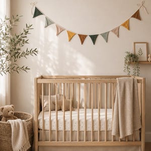 Könnte beinhalten: Ein hölzernes Kinderbett mit vertikalen Stäben und einer beigen Strickdecke, die über die Seite hängt. Im Inneren befinden sich ein beiges Kissen und ein sternförmiges Kissen. Darüber hängt eine Stoffwimpelkette in gedeckten Erdtönen.