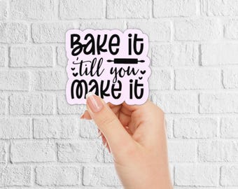 May include: A pink sticker with the phrase "Bake it till you make it" in black script. The sticker includes a rolling pin graphic. The sticker is held in front of a white brick wall.