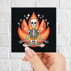 Op de afbeelding: Een skelet in een yogapose, zittend in een lotushouding met zijn armen uitgestrekt. Het skelet wordt omringd door een vurige oranje en gele vlam met witte sterren.