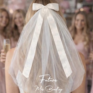 May include: A white bridal veil with a satin bow and ribbons. The sheer veil drapes down the back. Text on the veil reads "Future Mrs. Bradley". The background shows a group of women.