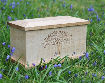 Puede incluir: Una caja de madera clara con un diseño de árbol tallado en la parte delantera. La caja está sentada sobre un lecho de flores azules.