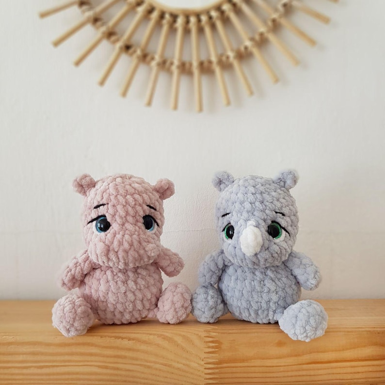 May include: Two crocheted stuffed animals, a pink hippopotamus and a gray rhinoceros, sitting on a wooden surface. Both animals have large, black eyes and are made with a textured yarn.