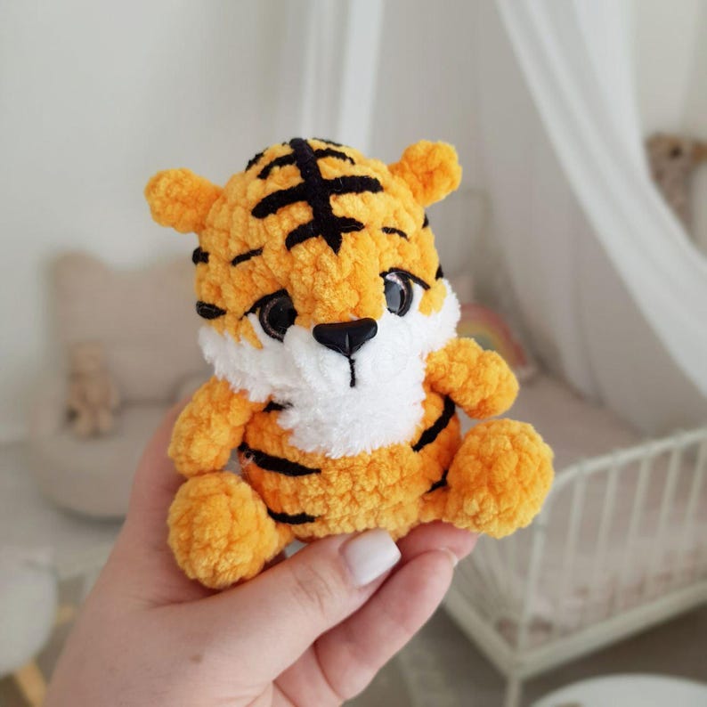 May include: A small crocheted tiger plush toy with orange fur, black stripes, and white facial details. The toy is being held in a person's hand.