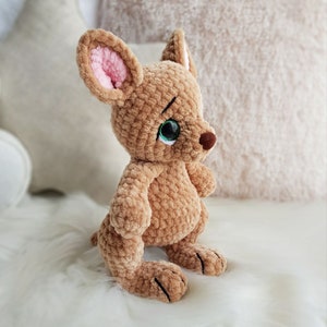 May include: A handmade, plush kangaroo toy in shades of brown with pink inner ears and green eyes. The soft, crocheted animal has a small tail and is sitting upright. The toy is on a white, fluffy surface.