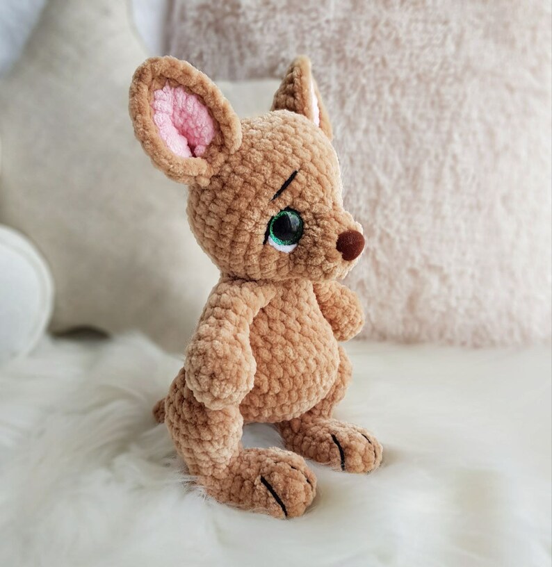 May include: A handmade, plush kangaroo toy in shades of brown with pink inner ears and green eyes. The soft, crocheted animal has a small tail and is sitting upright. The toy is on a white, fluffy surface.