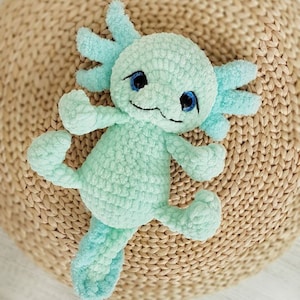 Pode incluir: Um brinquedo de pel&uacute;cia axolotl de croch&ecirc; com olhos azuis e um corpo verde claro. O brinquedo est&aacute; deitado em uma superf&iacute;cie de palha tecida.