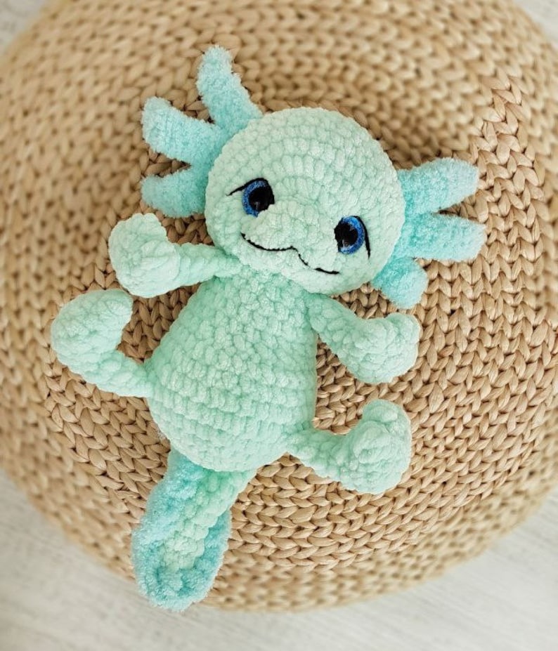 Pode incluir: Um brinquedo de pel&uacute;cia axolotl de croch&ecirc; com olhos azuis e um corpo verde claro. O brinquedo est&aacute; deitado em uma superf&iacute;cie de palha tecida.