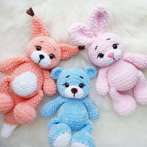 May include: Three crocheted stuffed toys: a pink bunny, a light blue bear, and an orange fox. The toys are all sitting on a white fluffy surface.