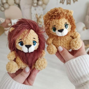 May include: Two crocheted lion plushies with brown and beige fur, blue eyes, and white noses. The lion on the left has a longer, reddish-brown mane.