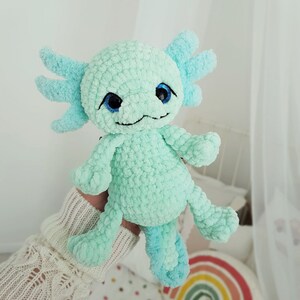 Pode incluir: Um brinquedo de pel&uacute;cia axolotl de croch&ecirc; com olhos azuis. O brinquedo &eacute; verde claro e tem uma superf&iacute;cie texturizada e felpuda.