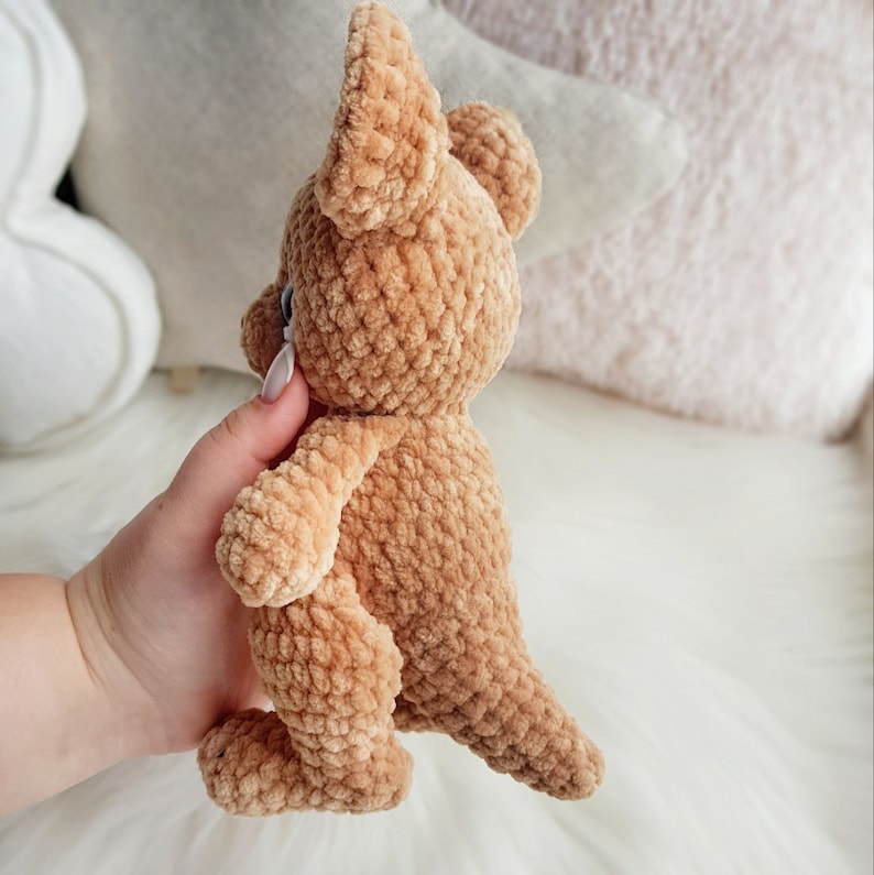 May include: A handmade, tan-coloured crochet kangaroo toy. The plush animal has large ears, a small tail, and is held by a hand. The toy is made of a soft, textured yarn and is set against a blurred background.