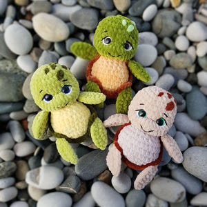May include: Three handmade crochet turtle plushies in shades of green, tan, and brown, with smiling faces and blue eyes. The turtles are arranged on a bed of smooth, gray and white stones, creating a whimsical scene.