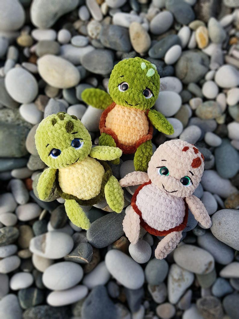 May include: Three handmade crochet turtle plushies in shades of green, tan, and brown, with smiling faces and blue eyes. The turtles are arranged on a bed of smooth, gray and white stones, creating a whimsical scene.