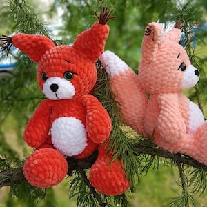 May include: Two crocheted plush fox toys, one red and one pink, sitting on a branch of a green tree. Both foxes have white accents on their bellies and black eyes.