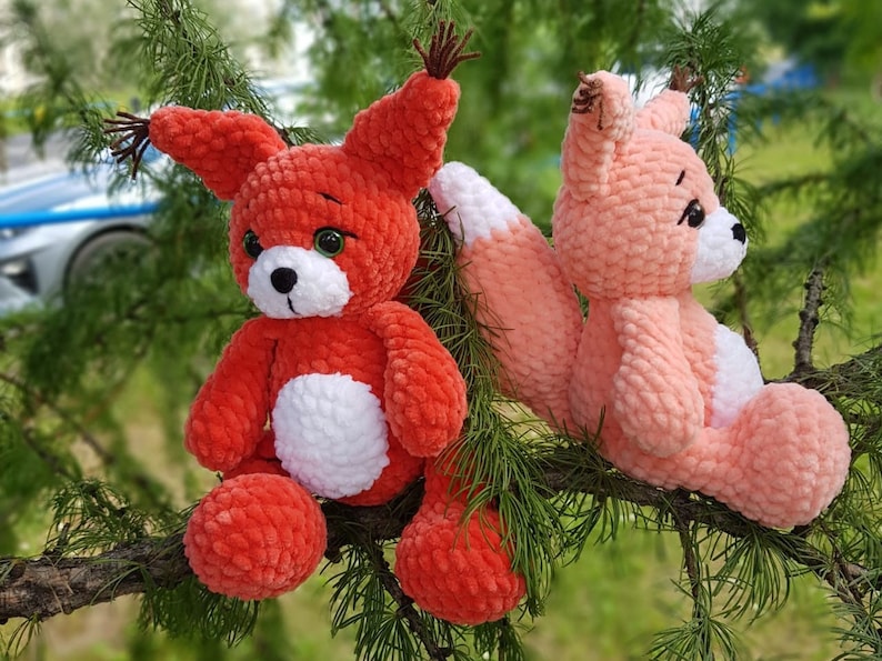 May include: Two crocheted plush fox toys, one red and one pink, sitting on a branch of a green tree. Both foxes have white accents on their bellies and black eyes.