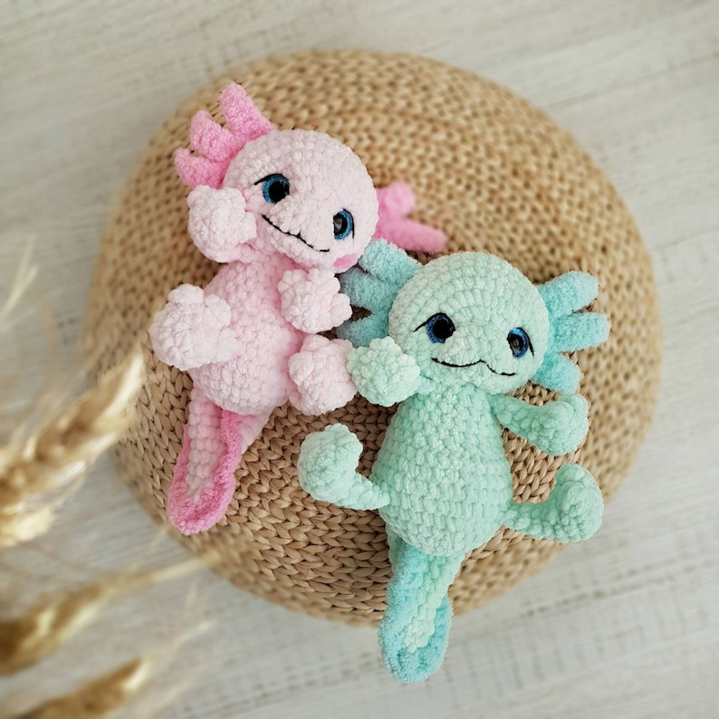 Pode incluir: Dois peluches de axolotl de croch&ecirc;, um rosa e outro azul claro, est&atilde;o sentados em um tapete de palha tran&ccedil;ado.