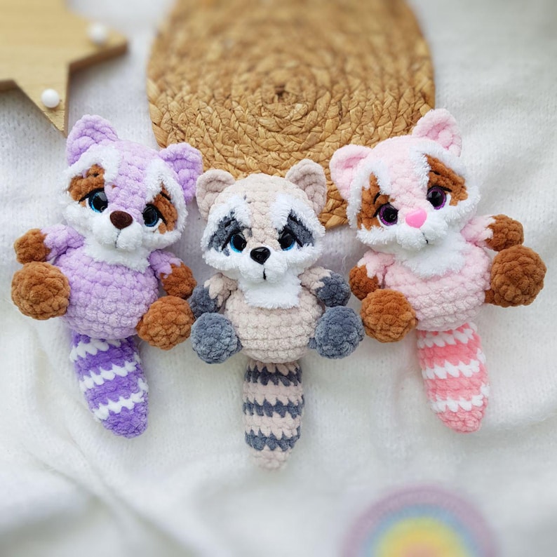 May include: Three handmade crochet raccoon plush toys in shades of lavender, tan, and pink. Each plush has a striped tail, brown paws, and a black mask around the eyes. The toys are displayed on a white surface.