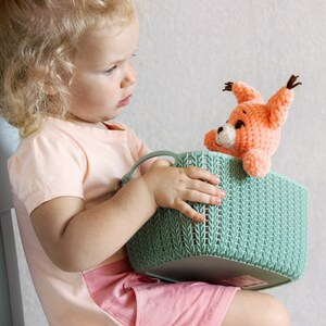 May include: A young child is holding a green woven basket with a crocheted orange fox toy inside. The child is wearing a pink top and a pink skirt.