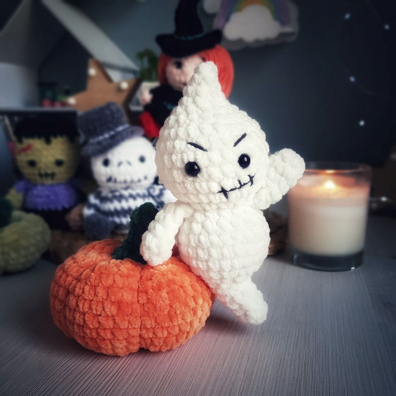 May include: A white crocheted ghost with a stitched-on mouth and eyes, holding a small orange crocheted pumpkin. The ghost is sitting on a wooden surface with other crocheted figures in the background.