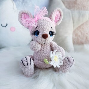 May include: A light pink crocheted mouse doll with a pink lace bow, blue eyes, and long eyelashes. The mouse holds a white daisy with a green stem. The doll is sitting on a white, fluffy surface.