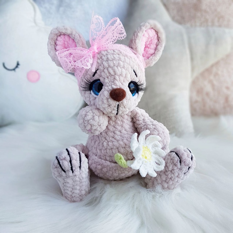 May include: A light pink crocheted mouse doll with a pink lace bow, blue eyes, and long eyelashes. The mouse holds a white daisy with a green stem. The doll is sitting on a white, fluffy surface.