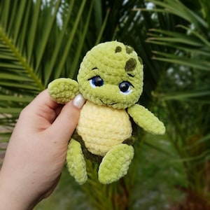 May include: A handmade, green and yellow crochet turtle with blue eyes. The turtle has a textured, plush appearance and is held against a backdrop of green foliage. The turtle's shell is yellow, and its head and limbs are green.