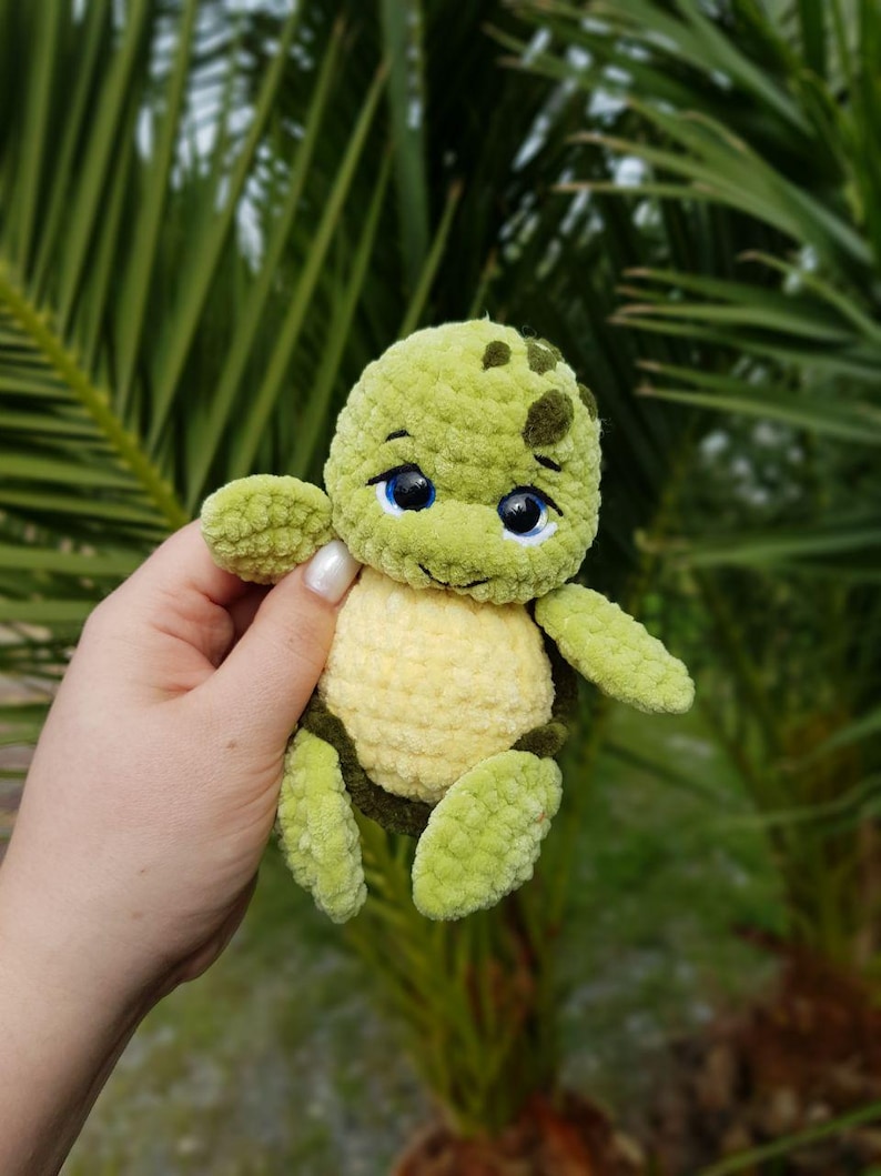 May include: A handmade, green and yellow crochet turtle with blue eyes. The turtle has a textured, plush appearance and is held against a backdrop of green foliage. The turtle's shell is yellow, and its head and limbs are green.