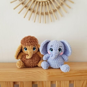 May include: Two crocheted stuffed elephants, one brown and one light blue, sitting on a wooden shelf. Both elephants have embroidered eyes and pink cheeks.