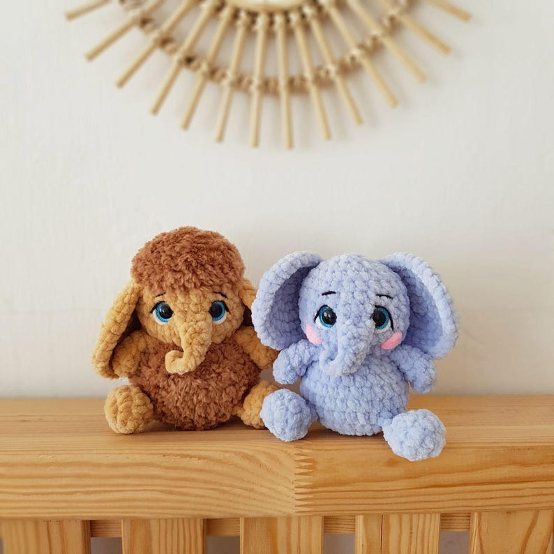 May include: Two crocheted stuffed elephants, one brown and one light blue, sitting on a wooden shelf. Both elephants have embroidered eyes and pink cheeks.
