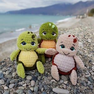 May include: Three handmade crochet turtle plushies in shades of green, yellow, and tan. Each turtle has a smiling face with blue or green eyes. The plushies are sitting on a bed of pebbles with a blurred ocean and mountain backdrop.
