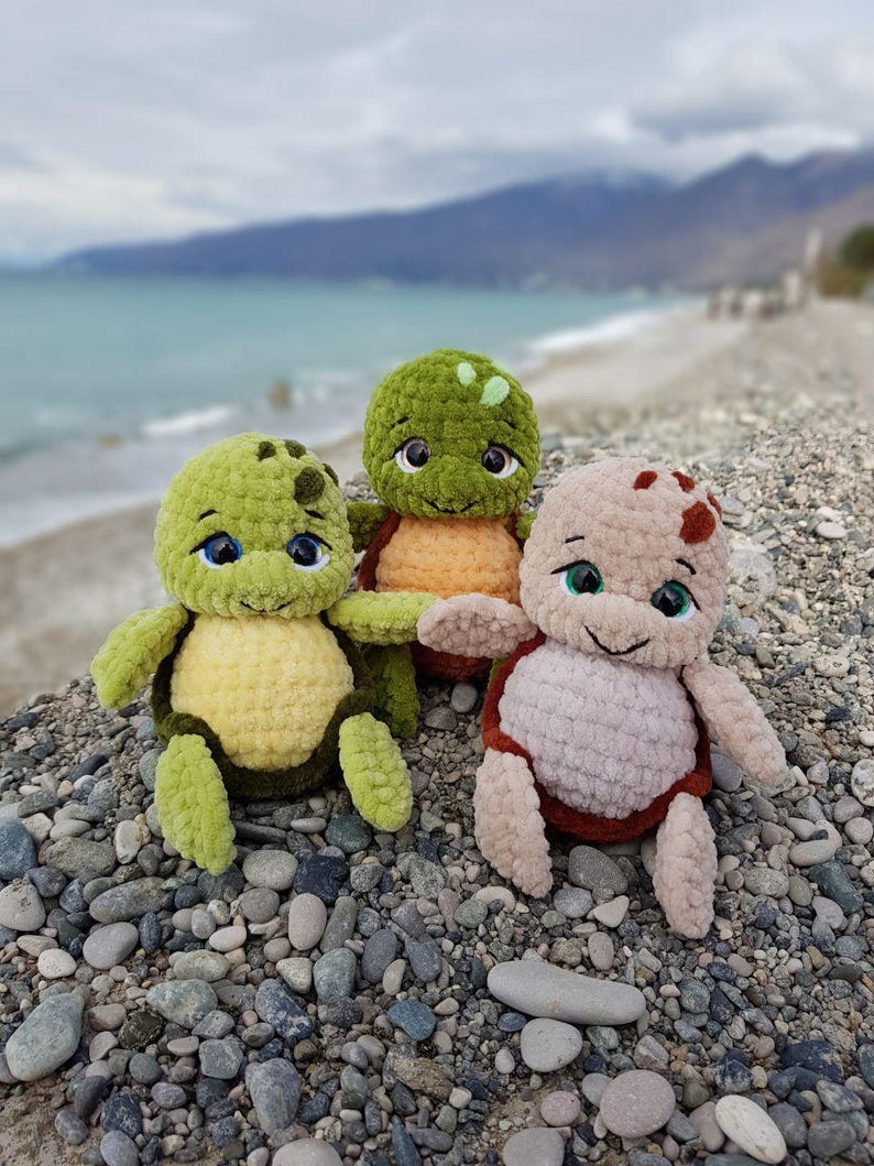 May include: Three handmade crochet turtle plushies in shades of green, yellow, and tan. Each turtle has a smiling face with blue or green eyes. The plushies are sitting on a bed of pebbles with a blurred ocean and mountain backdrop.
