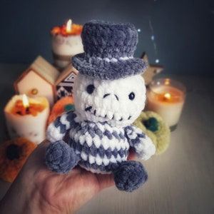 May include: A crocheted plush toy in the shape of a skeleton wearing a gray and white striped hat. The toy is sitting on a hand.