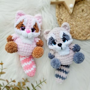 May include: Two handmade crochet raccoon plush toys. One is pink with brown accents and a pink and white striped tail. The other is gray with blue accents and a gray and white striped tail. Both have embroidered facial features.